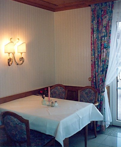 Stimmungsvolles Ambiente: Ein Tisch mit Stühlen vor einer tapzierten Wand, an der ein Leuchter hängt. Daneben sieht man ein Fenster mit Vorhängen.
